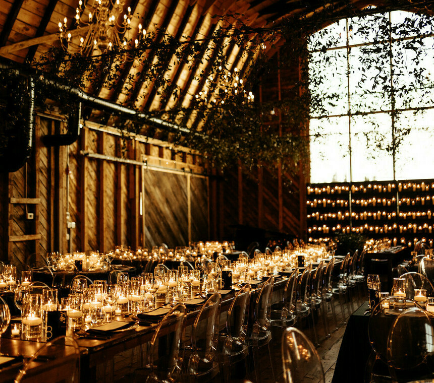 A luxurious barn wedding reception with round tables dressed in elegant place settings, crystal glassware, white floral centerpieces, glowing chandeliers, a wall of candles, and glass cylinder candles creating a warm, romantic ambiance. Tablescape rentals by Special Event Rentals.