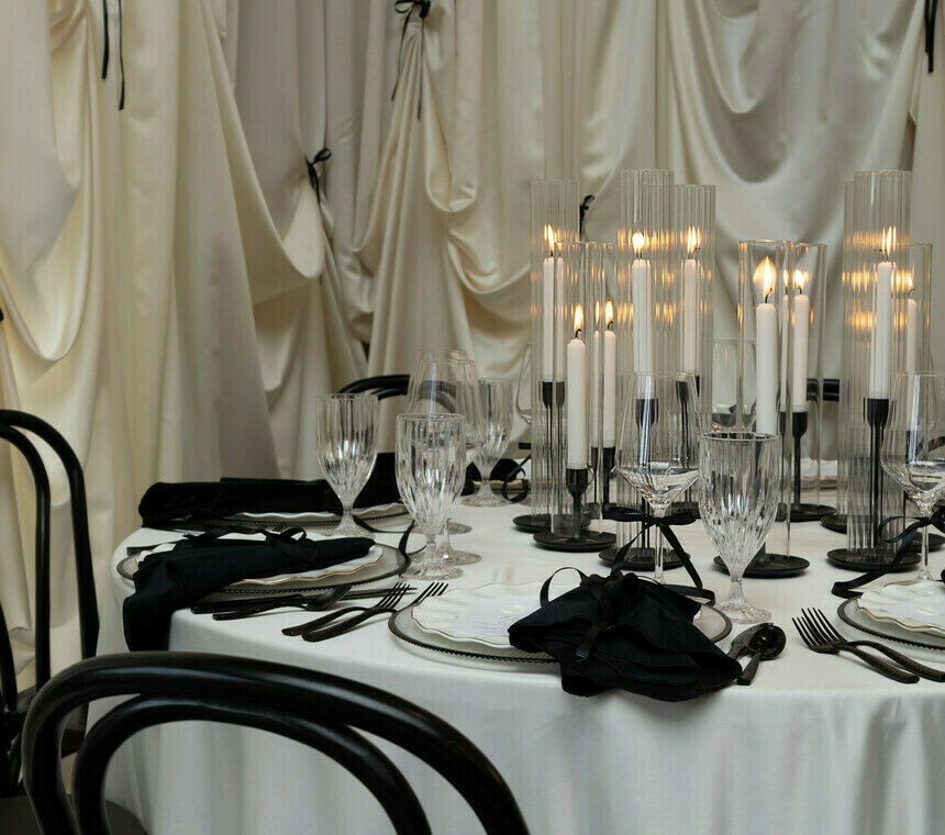 Elegant black and white table setting featuring a white tablecloth, black chairs, white plates, black napkins, refined cutlery, goblets, and candles with an off-white backdrop accented by black bow ties.