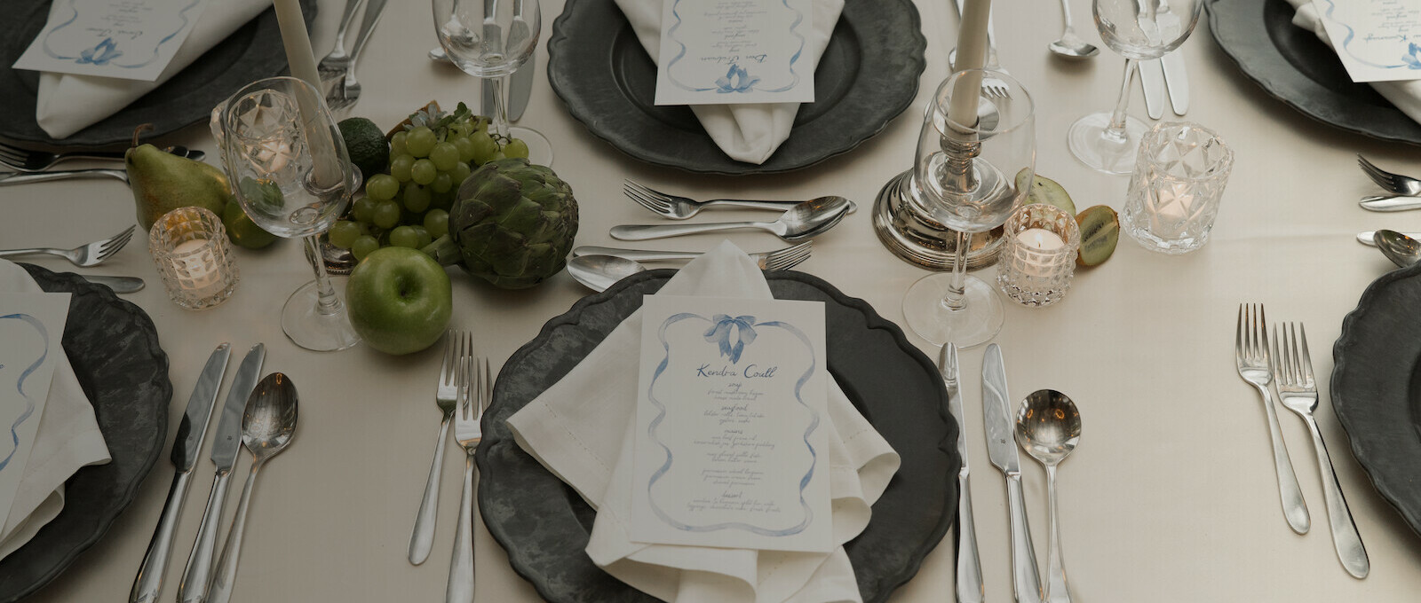 Elegant wedding table setting with black plates, white linens, candles, glassware, silver cutlery, and a natural fruit centerpiece featuring green apples, grapes, pears, and artichokes. Place cards rest on folded napkins at each setting, creating a sophisticated black-and-white tablescape.
