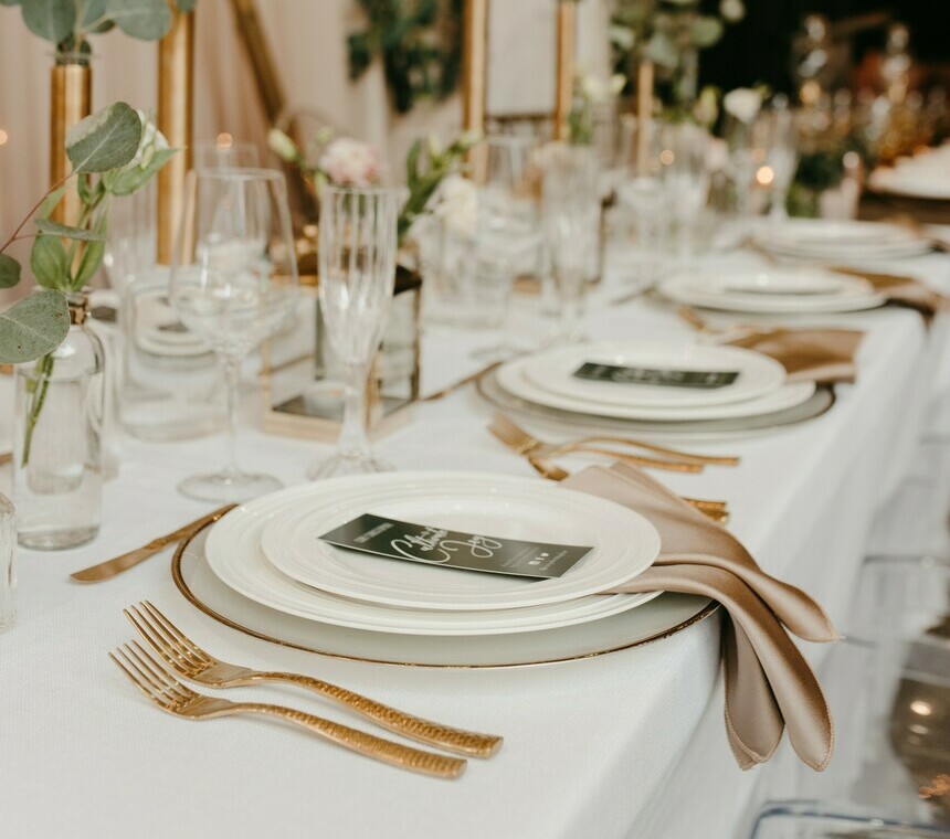 Elegant place setting featuring white plates, a knife, spoon, wine and champagne glasses on deep green linen, with a menu and candle creating a refined table setting.